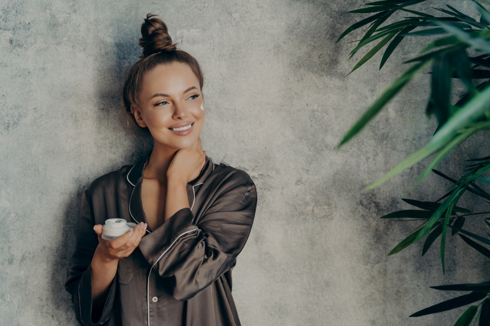 Young beautiful woman with glowing healthy skin broadly smiling while holding moisturizing face cream in her hand during morning cosmetic routine procedure at home, isolated over concrete wall. Young beautiful woman with glowing healthy skin broadly smiling while holding face cream