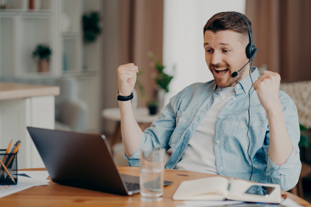 Overjoyed triumphing adult man clenches fists celebrates success looks attentively at laptop screen wears headset has corporate meeting online poses at desktop against blurred home background