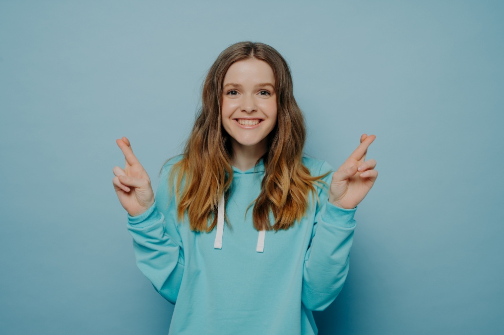Excited smiling young girl with wavy ombre hair keeping fingers crossed looking at camera wearing casual sweater posing isolated on light blue studio background. Body language concept. Happy young girl keeping fingers crossed making wish