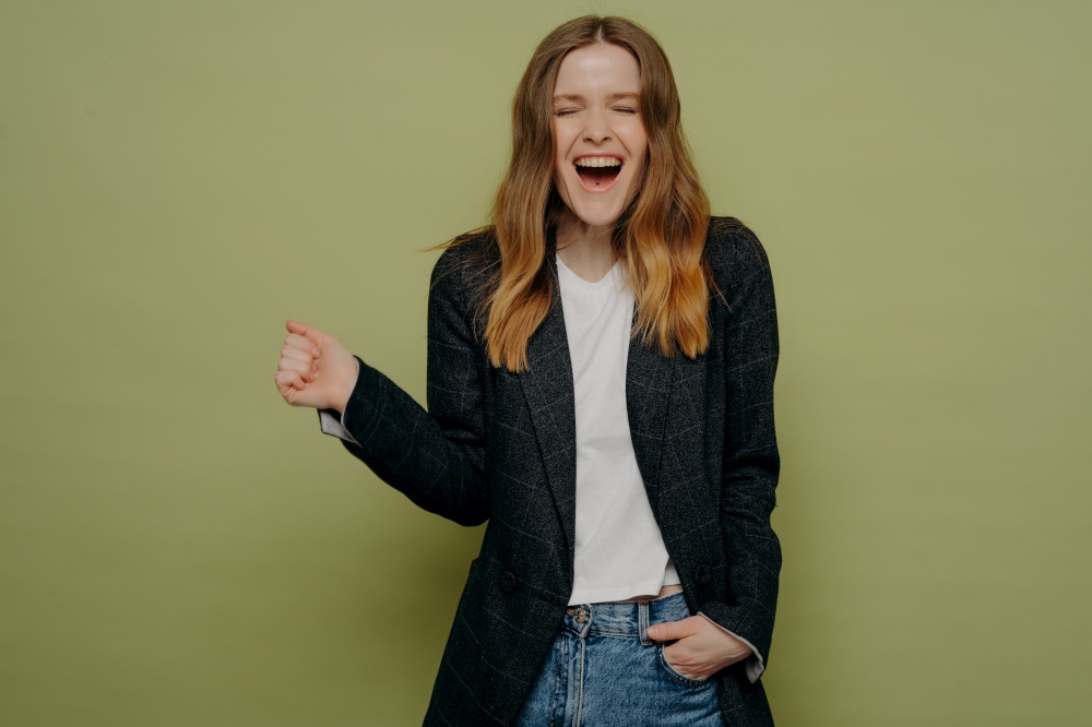 Excitement. Studio shot of surprised and happy young woman with wavy brown hair posing in dark formal jacket, white top and jeans on green background with one hand in pocket. Personal achievements. Excited young woman with closed eyes in formal jacket demonstrating happiness