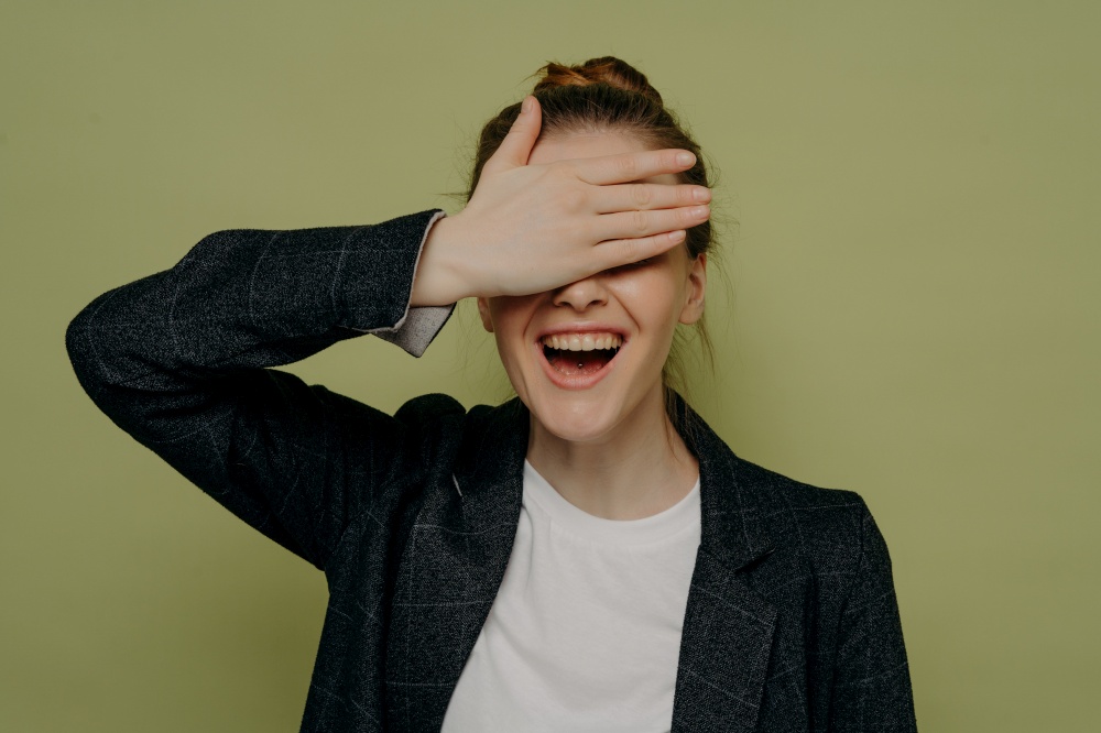 Laughing young female in casual clothes covering eyes with hand while waiting for surprise, smiling teenage girl keeping mouth opened with embarrassed expression, studio shot. Laughing young female in casual clothes covering eyes with hand while waiting for surprise