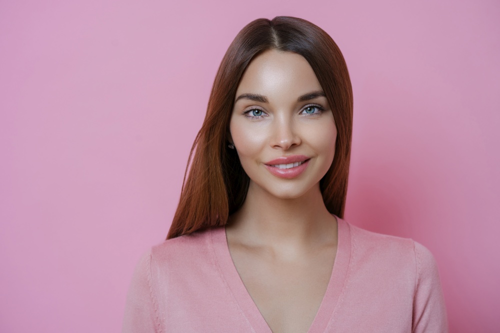 Close up portrait of attractive dark haired woman with brilliant smile, stands indoor against rosy background, has appealing look, isolated over pink background. Face model. Perfect fresh skin
