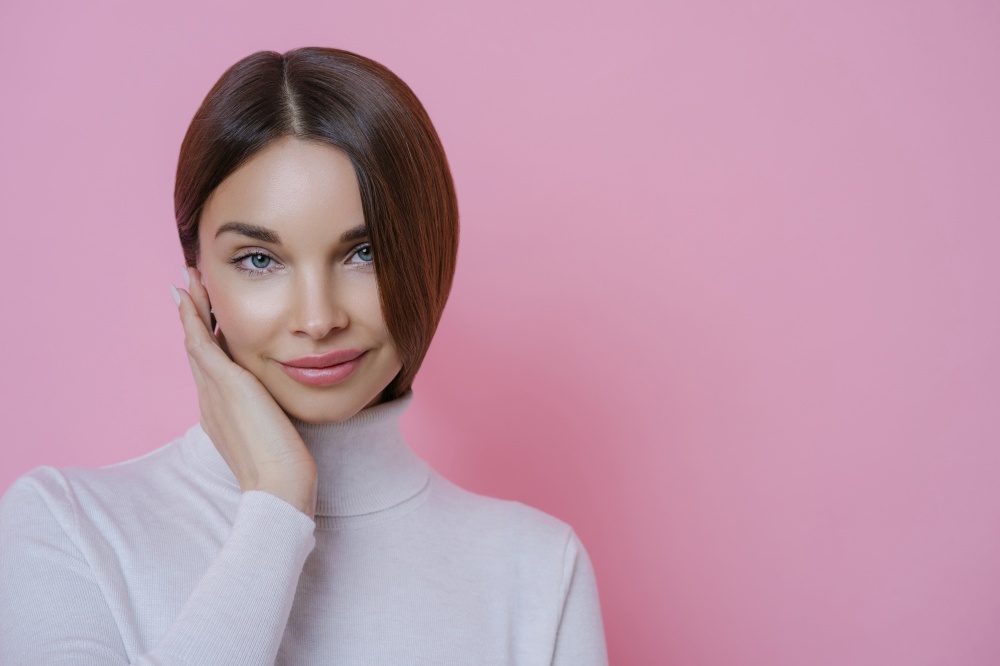 Isolated shot of pleasant looking dark haired young woman has well groomed face, dressed in turtleneck jumper, has healthy skin, isolated over rosy background. People, beauty, wellbeing concept