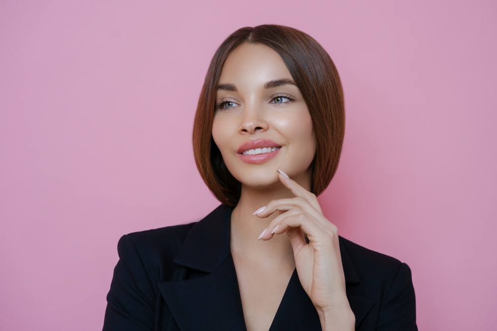 Portrait of satisfied lovely female employee looks away with dreamy cheerful expression, has manicure, dressed in black outfit, models against rosy background, thinks deeply about business affairs