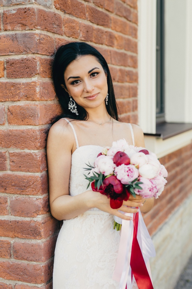 Attractive female bride wears wedding white dress, holds bouquet, stands near brick wall, has appealing appearance. Elegant young woman with make up, has charming smile celebrates her wedding