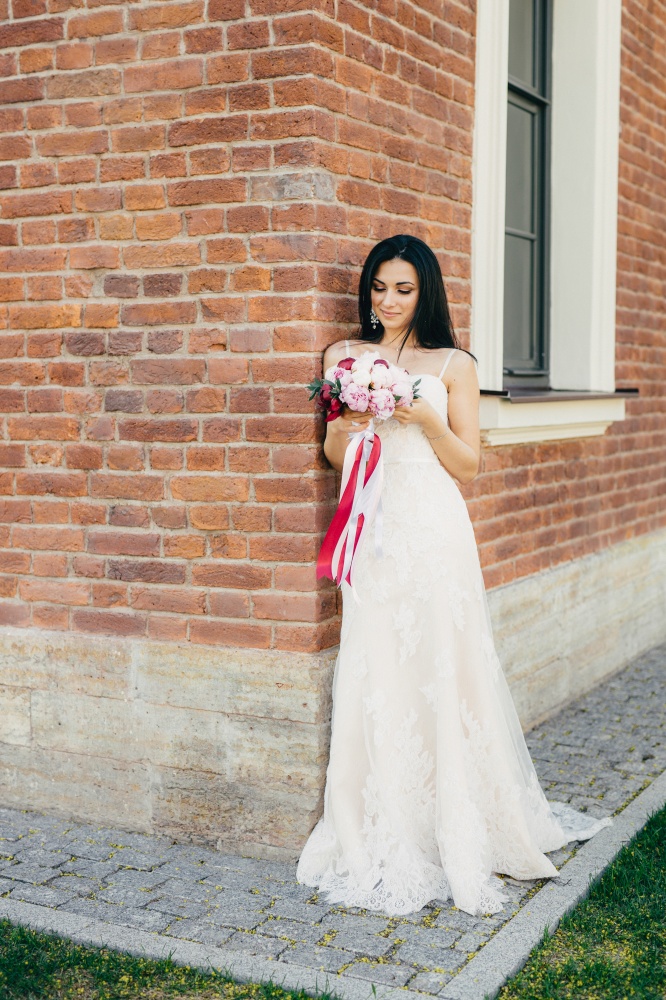 Thoughtful brunette female bride in beautiful wedding dress, keeps bouquete in hands, worries before special occasion in her life. stands near brick bilduing. Female model with perfect appearance