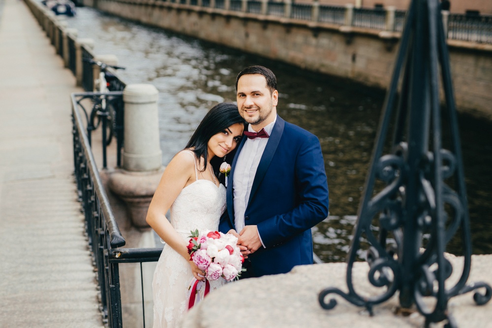 Affectionate couple in love celebrate their wedding, pose at camera as stand near bridge and river, have happy smiles. Glad positive married couple have romantic relationships. Wedding concept