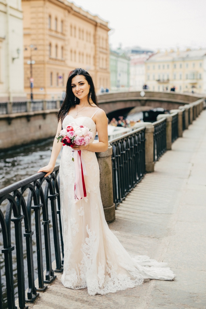 Vertical portrait of beautiful attractive female bride wears long white wedding dress, keeps bouquet , poses at camera as stands on bridge, has delightful expression. Happiness and beauty concept
