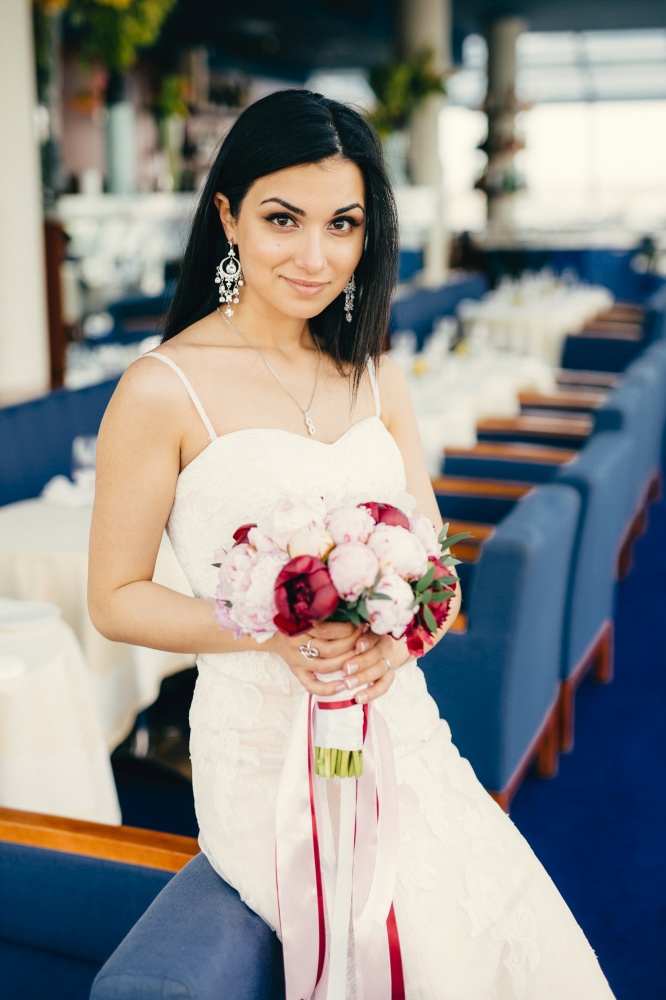 Pleased female model with appealing appearance wears white wedding dress, holds bouquet, being in festive hall, waits for bridegroom, looks directly into camera with charming shining eyes