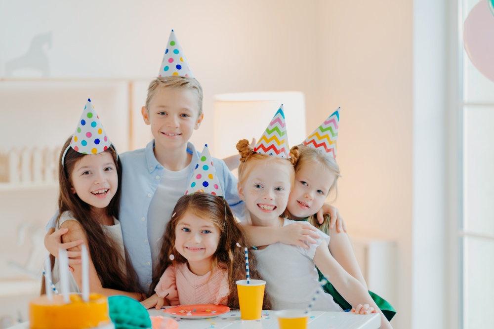 Group of adorable kids wear party hats, embrace and have fun, celebrate birthday, pose in decorated room, gather near festive table, have fun cuddle and look gladfully at camera