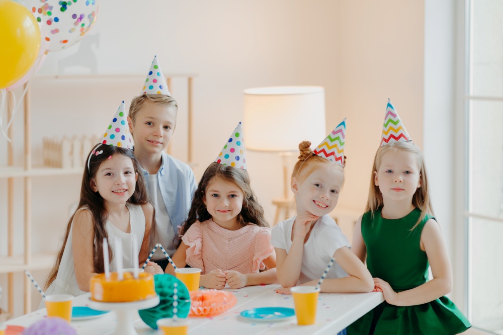 Children, celebration and birthday concept. Positive children have fun together at party, wear cone hats, eat delicious cake, play interesting games, being in good mood