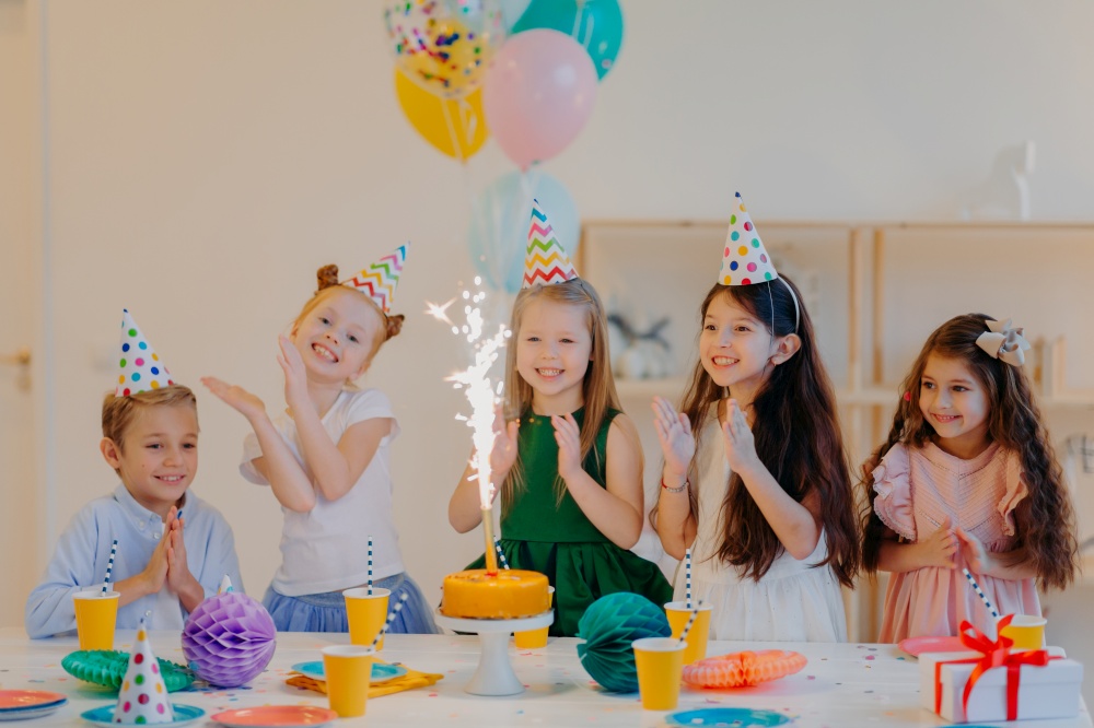 Children and holiday concept. Glad five friends look gladfully on cake with sparkle, celebrate birthday, wear party cone hats and hold air balloons, have happy expressions