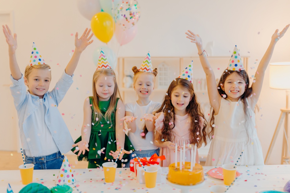 Horizontal shot of happy positive children catch confetti, celebrate birthday together, raise arms, have good mood, play together, stand near festive table with present box, cake, party hats