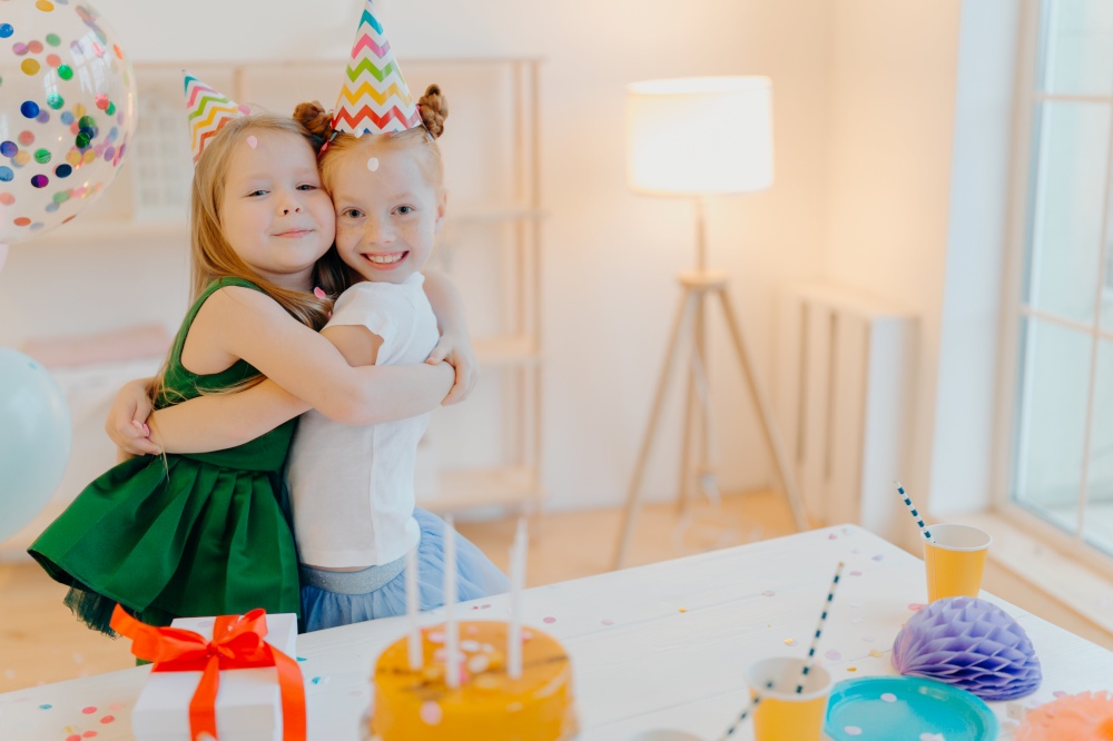 Friendly two girls embrace and have good relationship, stand near festive table with cake, celebrate birthday together, stand in living room. Glad female sisters enjoy holiday, special occasion
