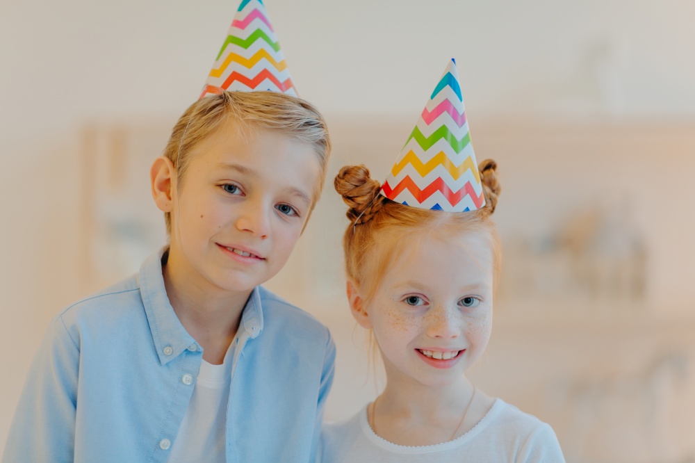 Portrait of happy ginger girl and her broghter stand closely to each other, wear party hats, come on friends birthday, look gladfully at camera, pose against blurred background. Childhood concept