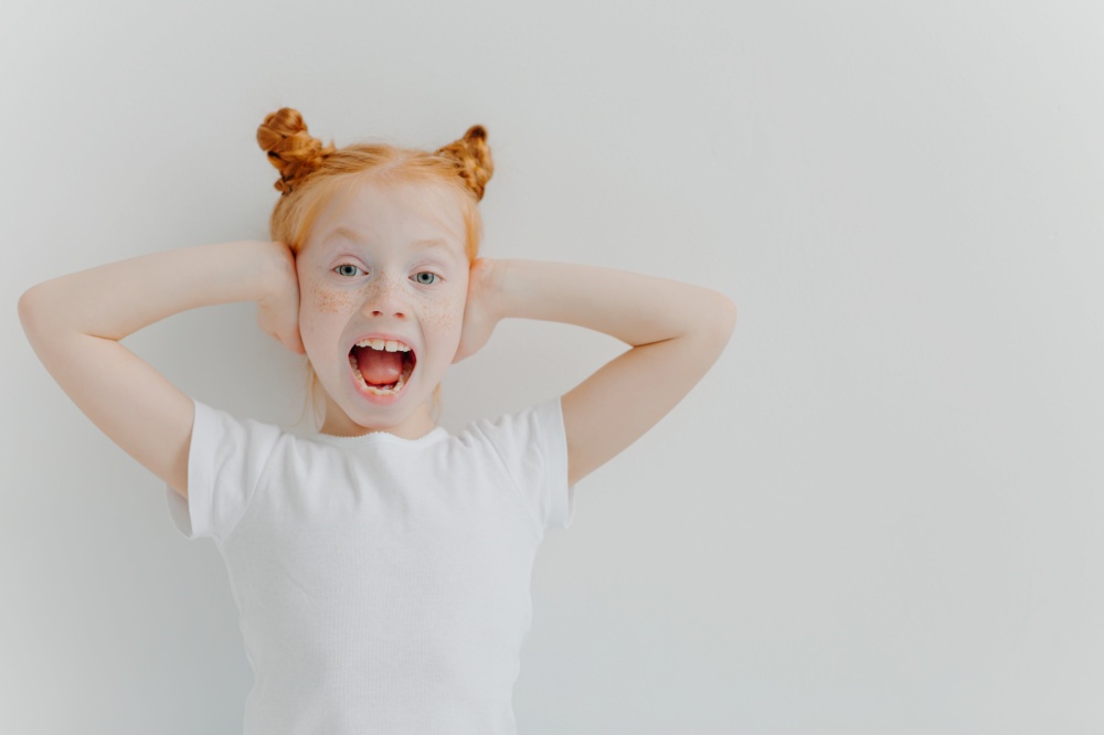 Emotional redhead girl keeps mouth widely opened, covers ears, excited by noisy atmosphere, wears white casual t shirt, reacts emotionally on something, has fun, isolated over white background