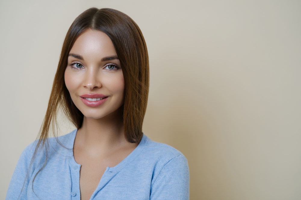 Portrait of lovely dark haired woman with pleasant appearance, full lips, wears casual blue jumper, poses against beige background, has calm expression sensual look healthy skin after facial treatment