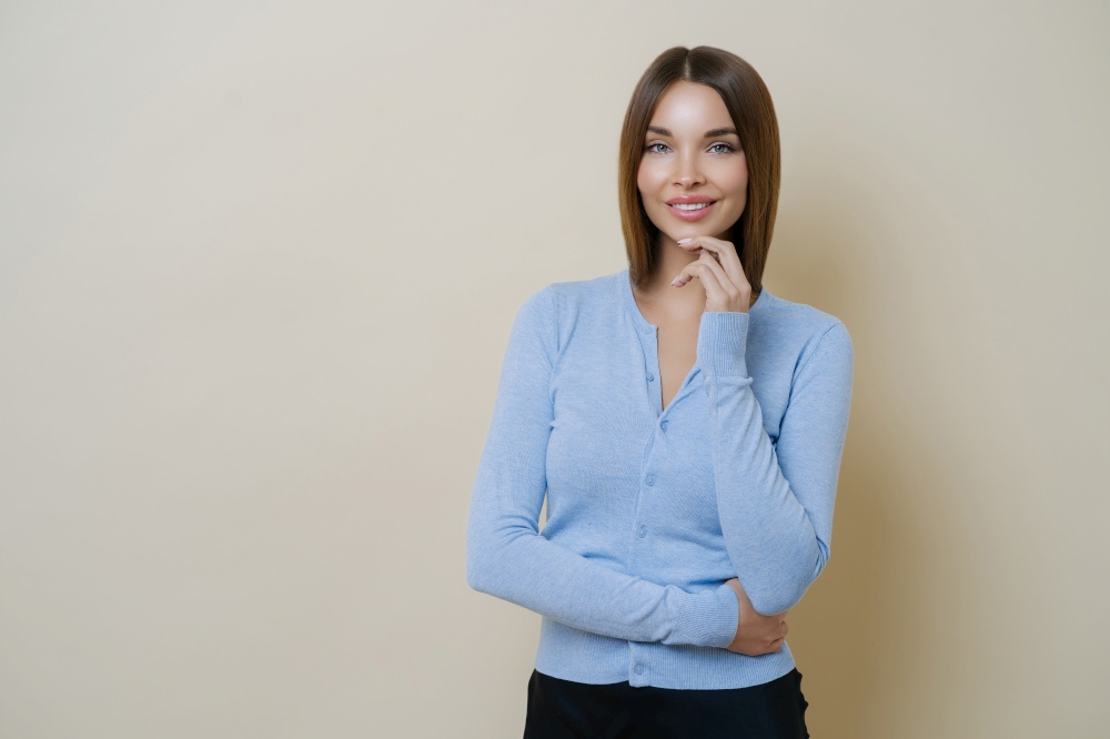 Half length shot of pretty slim woman dressed in casual blue jumper, keeps hand under chin, enjoys smooth perfect healthy skin, ready for work, isolated over beige background, blank space aside
