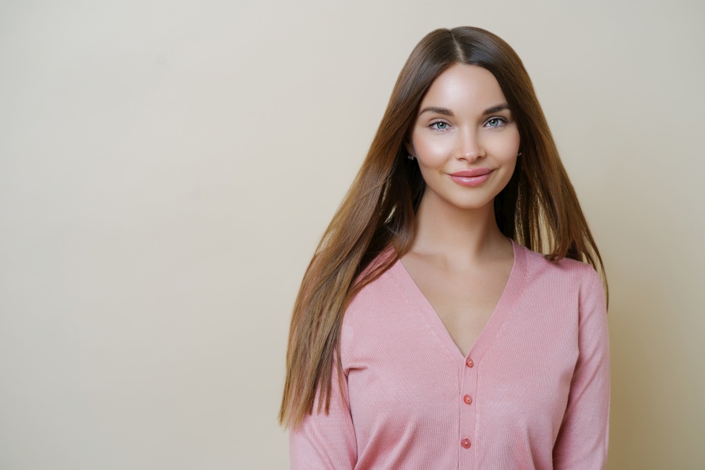 Horizontal portrait of charming European woman smiles gently at camera, has long straight hair, wears casual rosy sweater, going to have outdoor walk, stands against beige studio background.