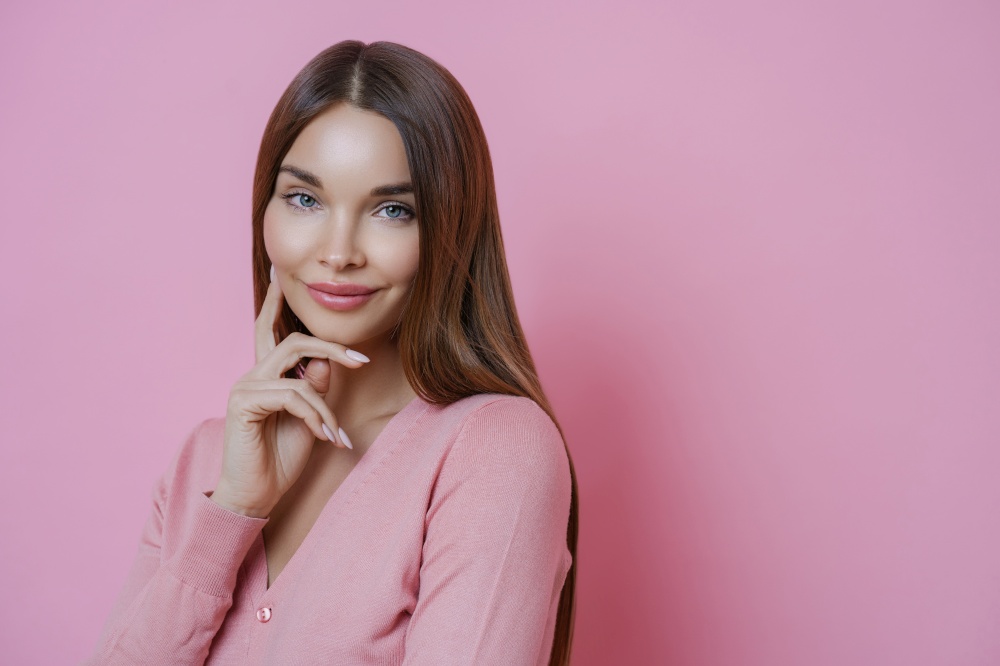 Young beautiful European woman touches face gently, looks with pleased expression at camera, has healthy perfect skin, listens friends advice, wears casual jumper, isolated over pink background