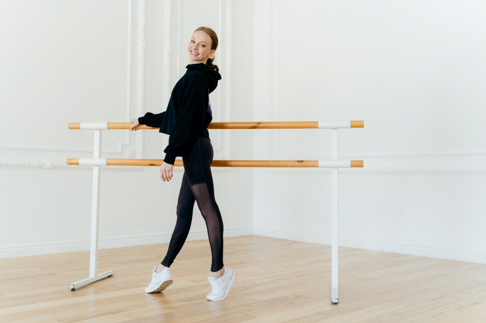 Slim beautiful red haired young woman stands sideways on tip toes, being in good body shape, trains every day, wears black sweatshirt, leggings and white sportshoes, keeps hand on ballet barre