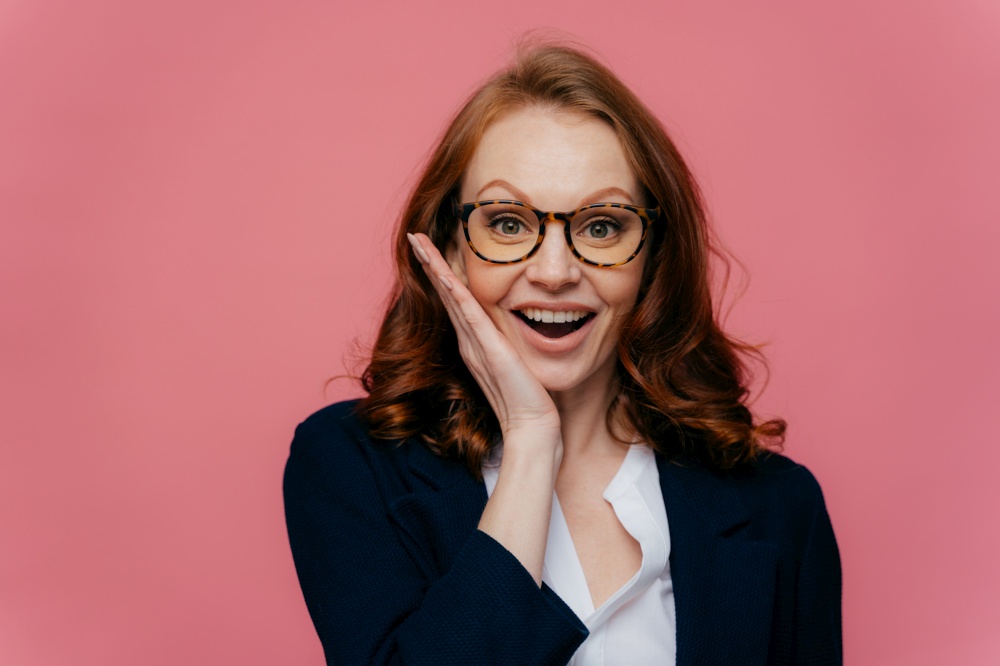 Happy female entreprenuer touches cheek gently, smiles broadly, wears transparent glasses, has ginger hair, dressed in formal apparel, satisfied to achieve success, isolated over pink background