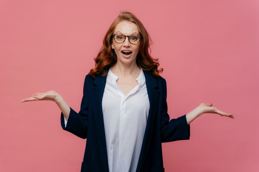 Half length shot of confused business lady raises palms, has hesitant facial expression, dressed in formal suit, spectacles, expresses bewilderment, isolated over pink background. People and doubt