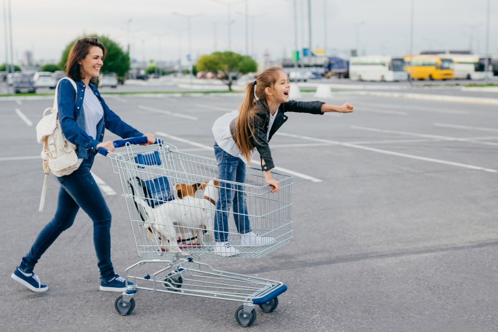 Photo of happy small girl with her mother and pets stands in cart, make shopping during weekend, enjoy free time, return home with new purchases. Family, leisure, animals and purchsing concept