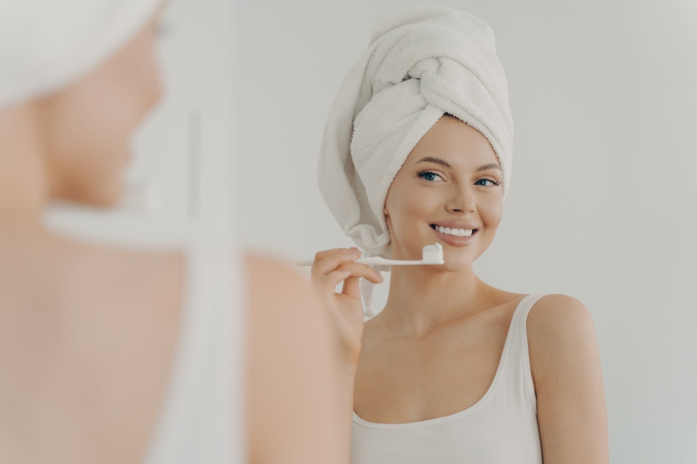 Beautiful young woman with healthy perfect smile brushing teeth and looking in mirror, attractive young female wearing white bath towel on head standing in bathroom at home. Oral hygiene concept. Beautiful young woman with healthy perfect smile brushing teeth and looking in mirror