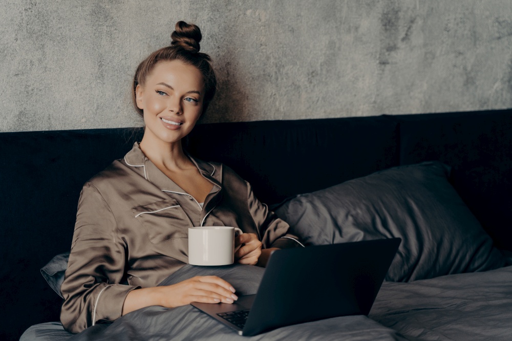 Lazy relaxed morning time. Smiling beautiful young female lying in bed in satin pajama with cup of coffee in her hand while checking last news on internet online on laptop, enjoying free time at home. Smiling beautiful young female using laptop while lying in bed with cup of coffee