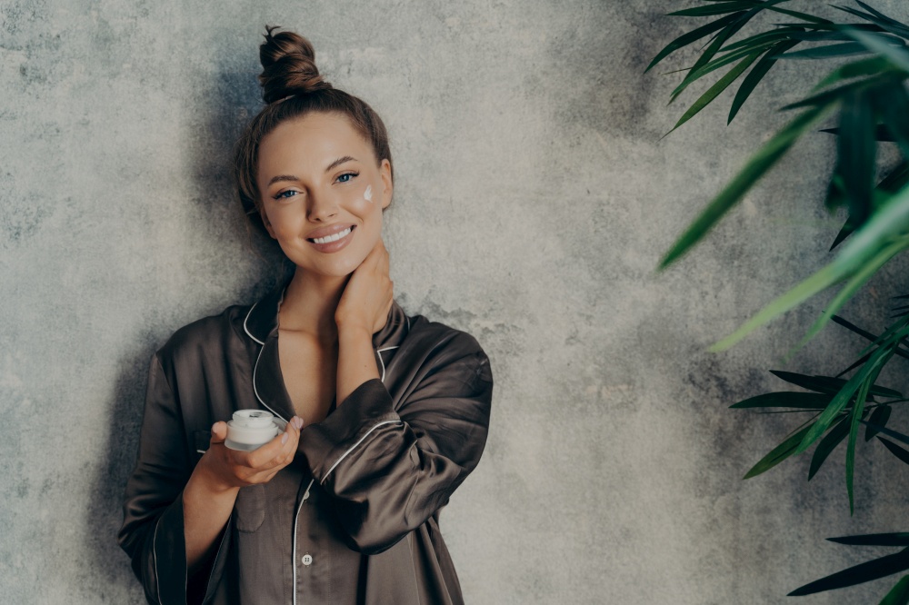 Image of cheerful woman in silk pajama smiling while applying face cream, posing with jar of moisturizing lotion in her hand, isolated over concrete wall background. Beauty and skin care concept. Beautiful caucasian woman in silk pajama with face cream smiling at camera
