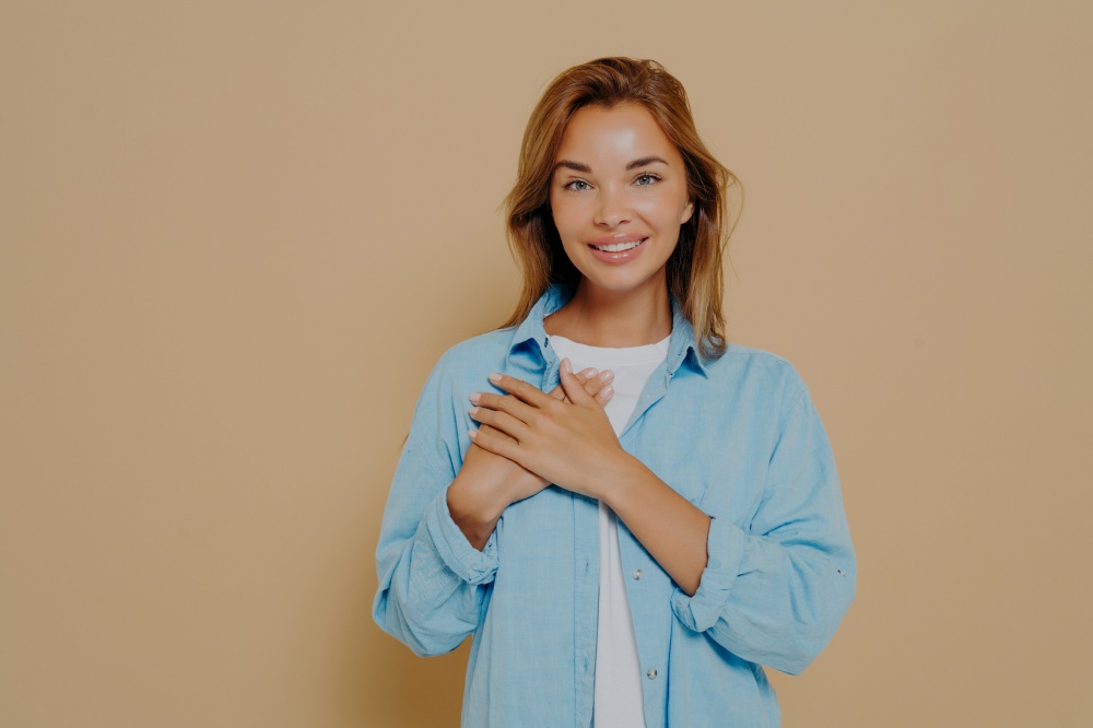 Delighted beautiful woman demonstrating her touched lovely mood by holding hands near heart, looking at camera with love and gratitude while posing in casual outfit on beige background. Smiling woman holding hands near heart on beige background