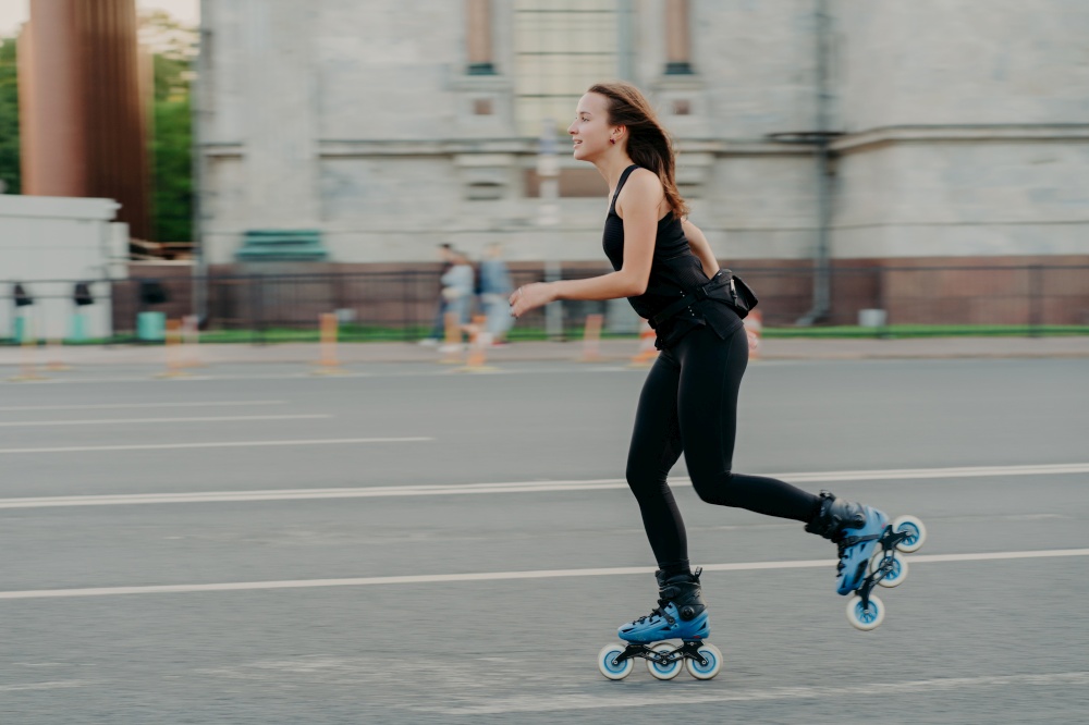 Summer rollerblading and fitness concept. Fit young dark haired woman fond of rollerskating exercises all body poses on road along asphalt during sunny day wears comfortable clothes has sporty figure