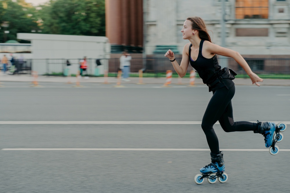Horizontal shot of sporty energized woman enjoys rolleblading being photographed in motion poses on road against blurred street background has regular fitness activities for keeping fit and healthy