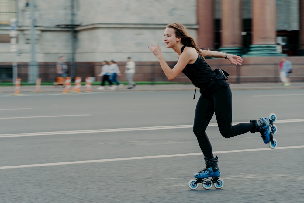 Holidays and active lifestyle concept. Slim healthy European woman rollerskates on high speed photogaphed in motion dressed in black clothing poses against blurred street background on road.