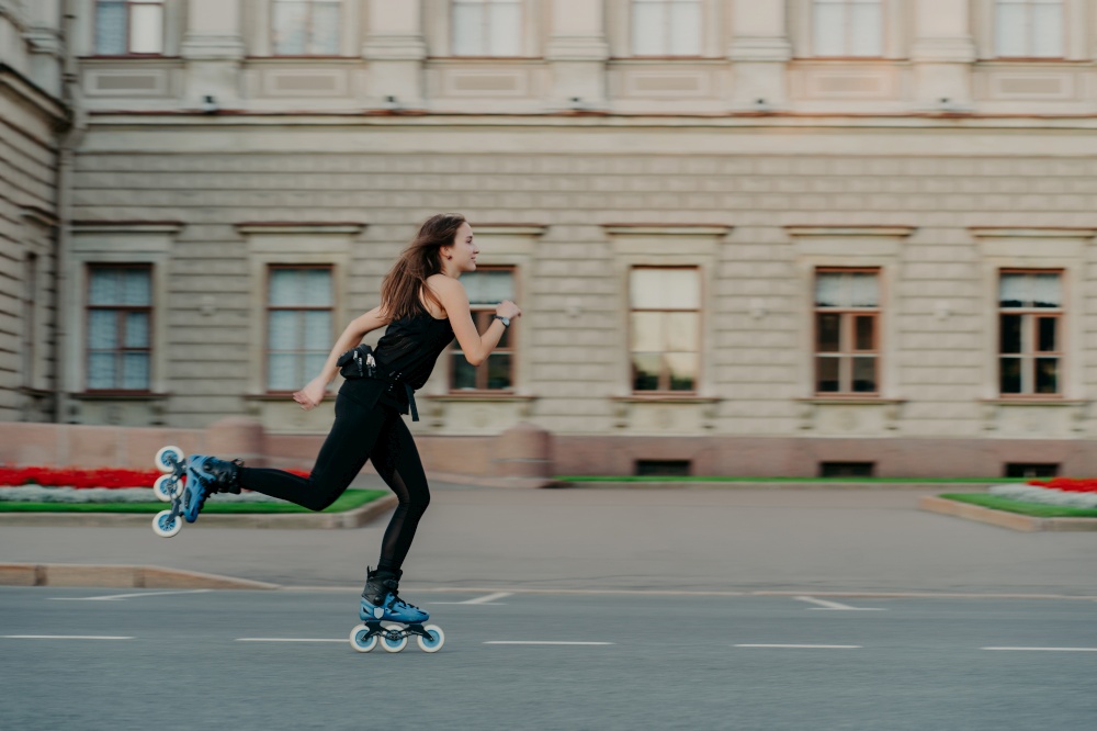 Outdoor physical activities. Healthy young woman has slim fit figure leads active lifestyle spends free time outside rides on rollerblades poses alone in open air against building background