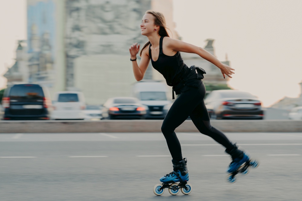 Young dark haired slim healthy woman has recration time enjoys rolleblading on speed moves fast has cheerful expression wears black t shirt and leggings. Active rest and sport training concept
