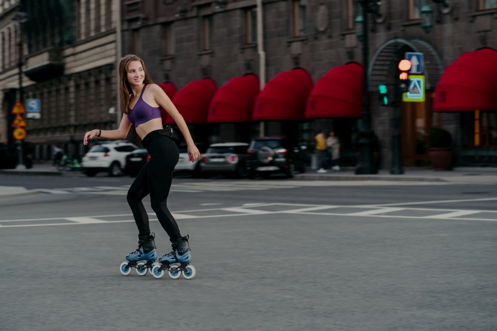 Outdoor shot of active slim woman dressed in cropped top and leggings rollerblades through city enjoys excellent physical workout burns calories gets energy relieves stress learns new skills