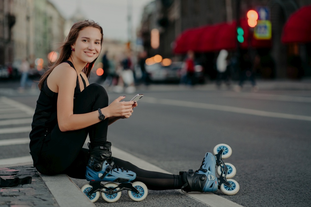 People outdoor activities and recreation concept. Horizontal shot of active slim woman being in good physical shape rides rollerblades uses smartphone sends text messages online poses outside