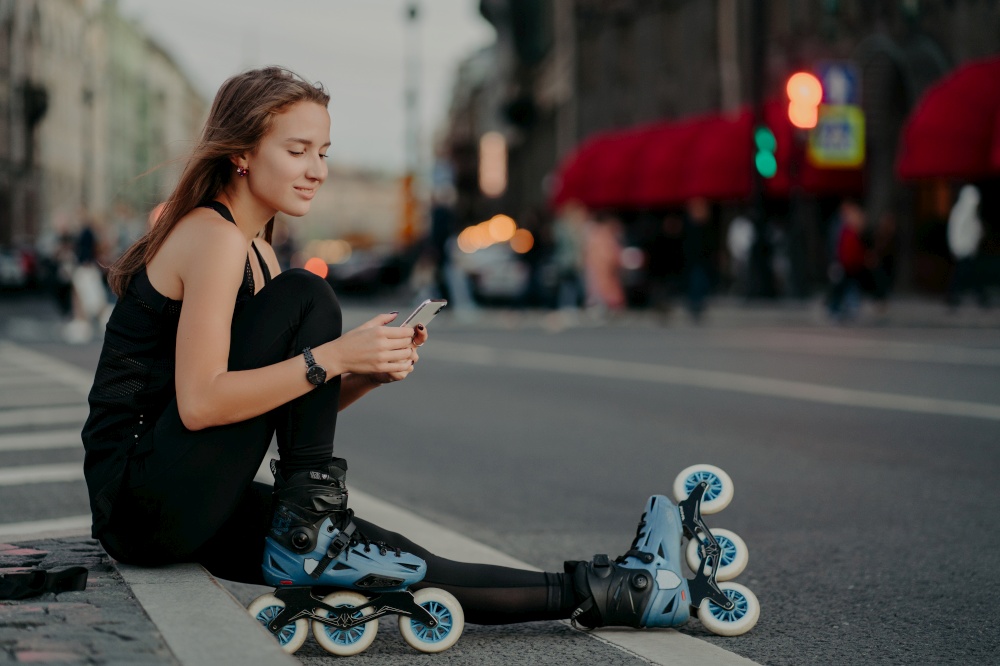 Outdoor shot of sporty active young woman dressed in active wear wears rollerblades checks message content on smartphone poses against blurred city background on asphalt leads healthy lifestyle