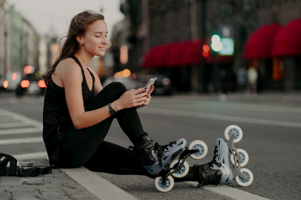 Horizontal shot of active sporty woman dressed in sportsclothes wears rollerblades focused at smartphone enjoys inline skating outdoors leads active lifestyle. Rollerskating as hobby concept