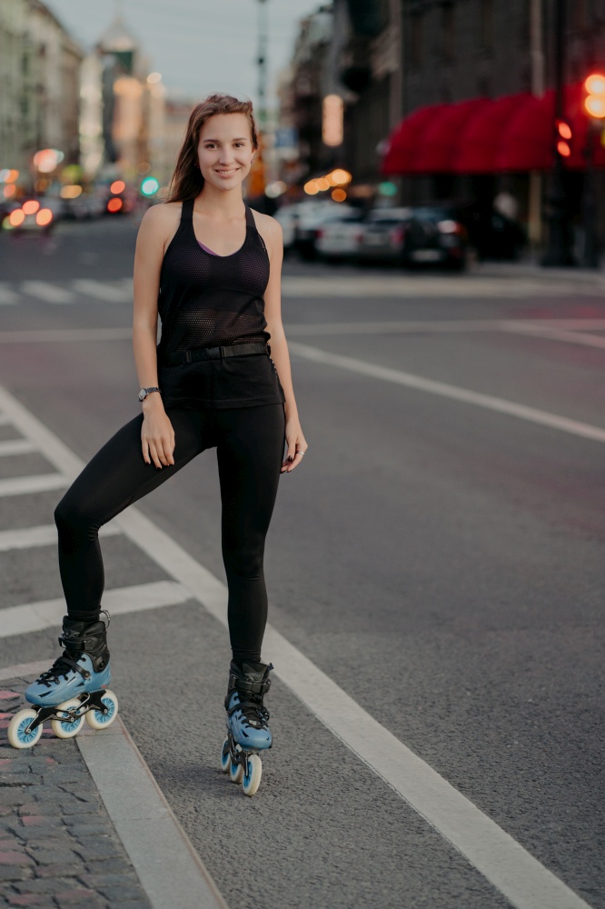 Full length shot of active young woman being in good physical shape dressed in black activewear enjoys rollerblading during good summer day poses on road against city background. Recreation.
