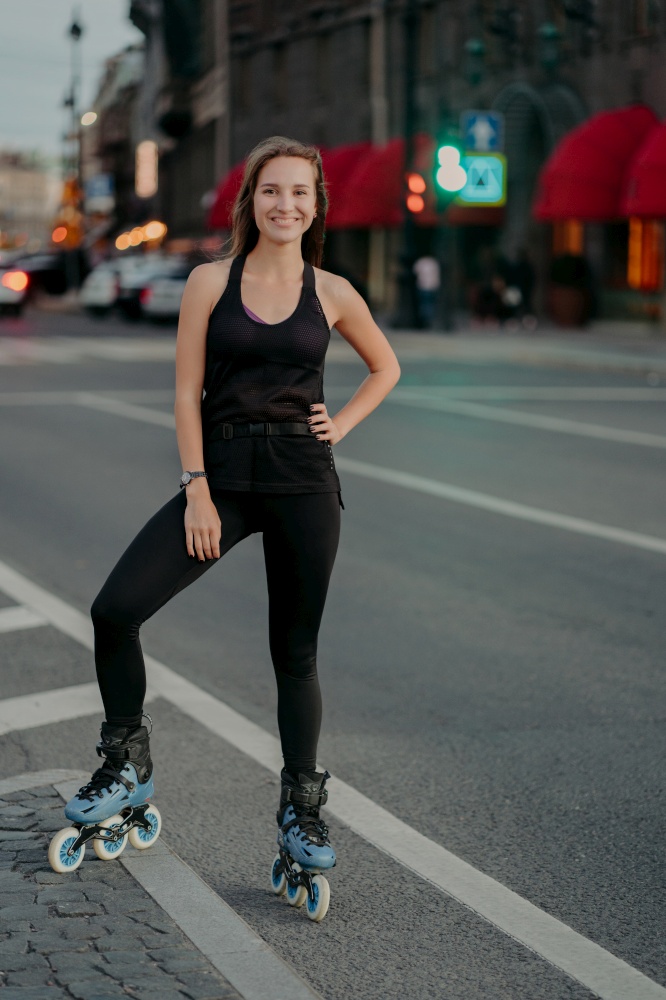 Pleased Eurpean woman keeps hand on waist smiles pleasantly leads active lifestyle rides rollerskates dressed in black clothing poses against blurred busy city background. Extreme sport concept