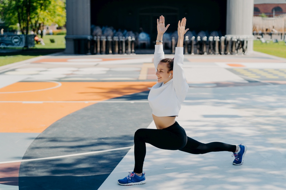 Flexible fit sportswoman does sport exercises outdoors keeps arms raised has glad expression wears hoodies leggings and trainers poses on stadium in urban place listens favorite audio track.
