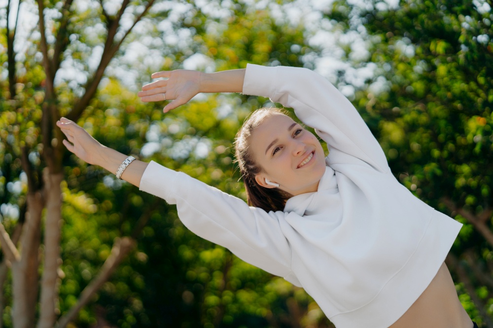 Happy active young woman stretches arms leans aside smiles positively enjoys exercising in fresh air wears white sweatshirt has regular trainings or workout poses outdoors. Wellness recreation concept