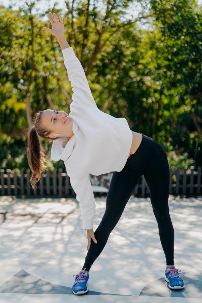 Slim healthy woman exercises outdoor keeps arm raised stretches body before running enjoys physical activity dressed in active wear breathes fresh air listens music from playlist. Sport concept
