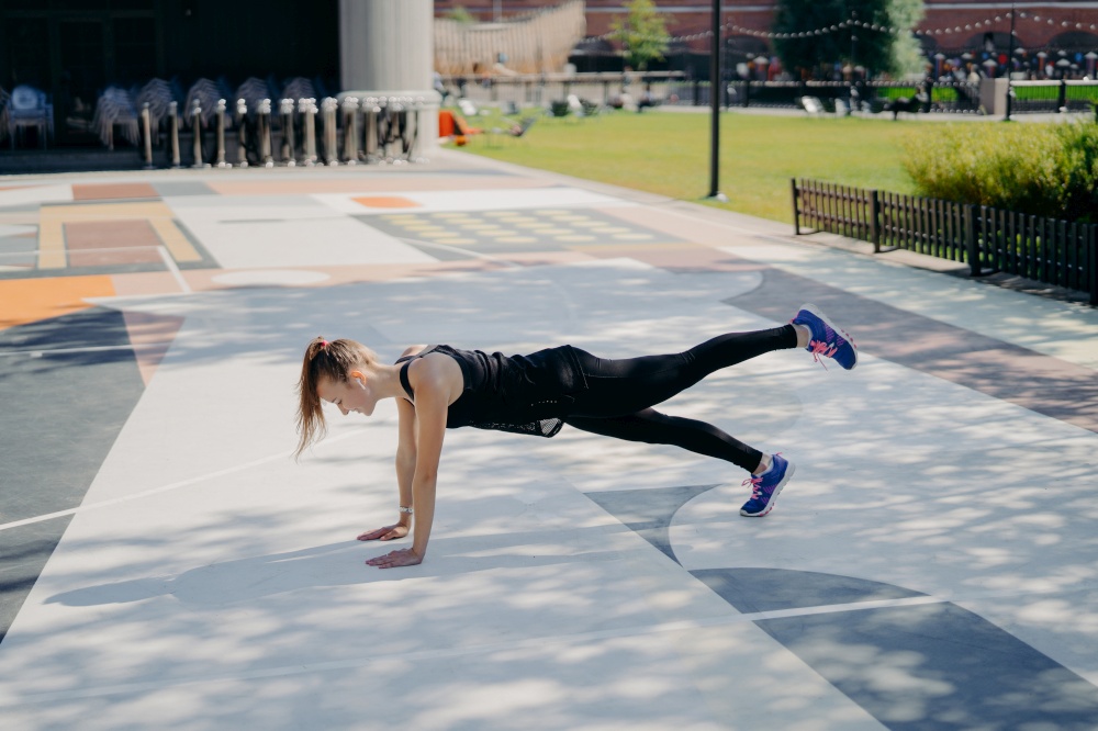 Sporty slim young woman does planking exercise raises leg demonstrates her determination wears sportsclothes and sneakers trains outdoors listens music in earphones. Healthy lifestyle concept