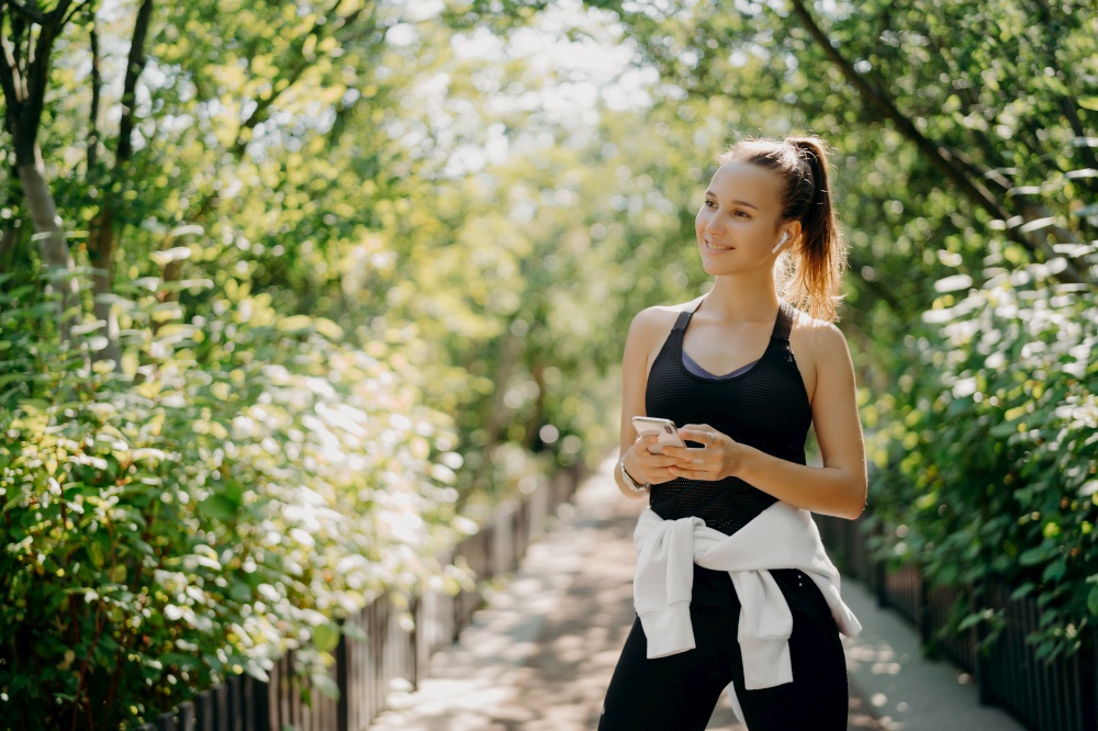 Horizontal shot of active sporty woman uses smartphone for checking results after jogging dressed in active wear enjoys warm sunny day listens music via earphones. Healthy lifestyle concept.