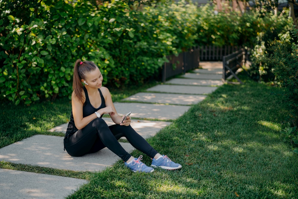 Sporty female blogger talks with followers online talks about healthy lifestyle and fitness training shares media in networks uses wireless earphones dressed in activewear poses against green grass