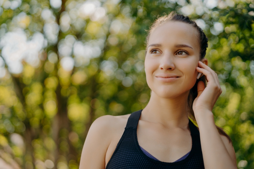 Healthy lifestyle concept. Outdoor shot of sportswoman enjoys favorite playlist in earbuds while training in park has gentle smile concentrated into distance poses over blurred nature background